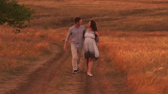 Happy Pregnant Woman and Her Husband are Walking Along a Country Road at Sunset