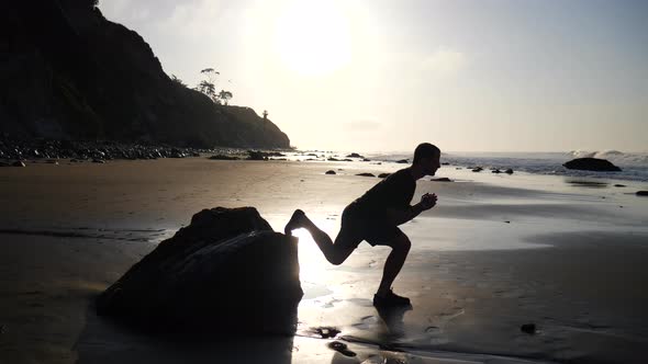 A strong muscular man doing leg lunges for a morning fitness workout at sunrise on a beach in Santa