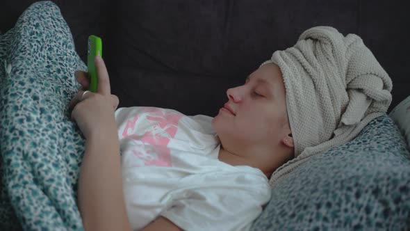 Young Woman with Towel on Her Head is Lying in a Straightened Bed Under Blanket in Pajamas and