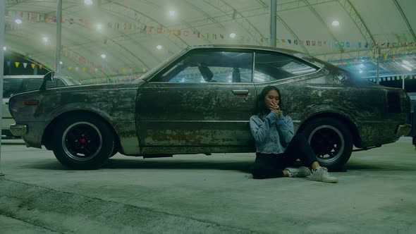 a Sad Thai Girl Sits Smoking Leaning Against a Rusty Car at Night