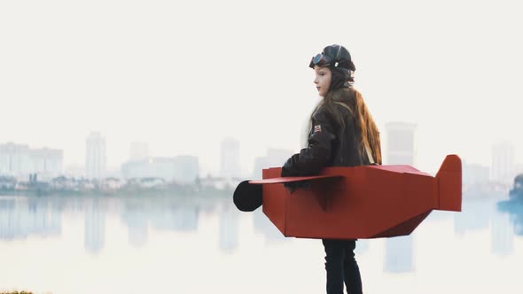 Happy Little Pilot Girl Standing at Calm Lake City Panorama in Cardboard Plane Playing Aviator