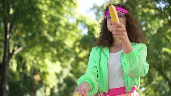 Carefree Laughing Woman in Retro Outfit Imitating Gun Shooting with Bananas Outdoors in Slow Motion