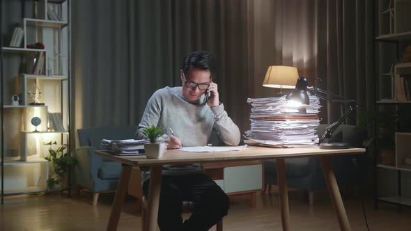 Asian Man Talking On Phone And Taking Note While Working Hard With Documents At Home