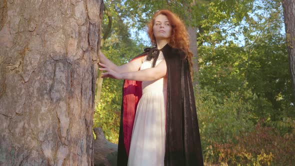 Beautiful Caucasian Girl with Red Hair in Long White Dress and Black Gown with Red Lining Standing