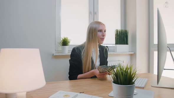 Woman Celebrating Success in Office. Elegant Blond Female Sitting at Workplace Holding in Hands
