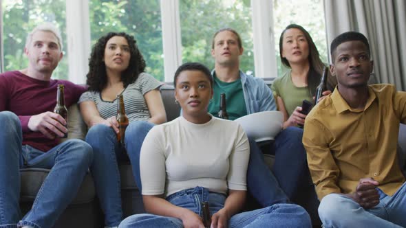 Group of diverse young people holding beers cheering while watch tv at home