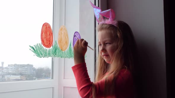 Cute little girl in bunny ears painting Easter eggs with green spring grass on window at home