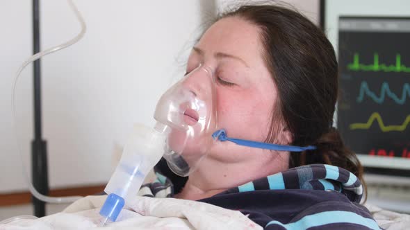 Woman with Eyes Closed in Oxygen Mask in the Background of a Monitor with Vital Indicators