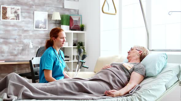 Nurse in Retirement Home Talking with an Old Lady Lying in Hospital Bed