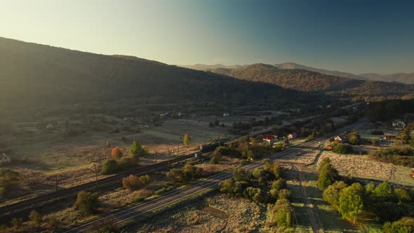 Aerial View Asphalt Road and Locomotive Moving By Railway at Sunny Sunrise in Country Side in