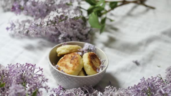 A Bouquet of Lilac Flowers Near a Plate with Cheese Cakes on a White Background