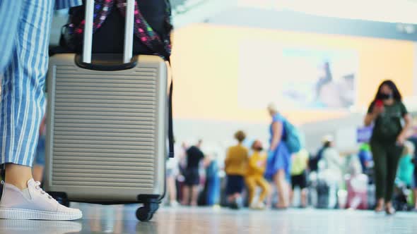 Close-up of Walking Legs in Sneakers, with Suitcase on Wheels, Through Airport Lobby, Against