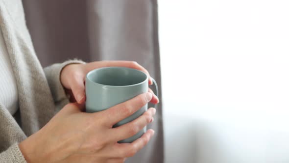 The Girl Drinks Hot Tea or Coffee From a Mug, Standing at the Window. Steam Comes From the Cup