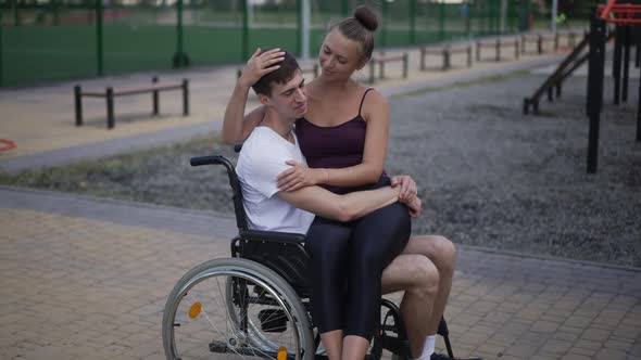 Loving Smiling Woman Stroking Hair of Young Man in Wheelchair Sitting on Lap