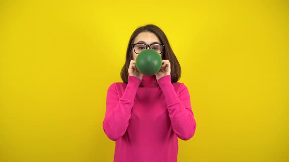 A Young Woman Inflates a Green Balloon with Her Mouth on a Yellow Background. Girl in a Pink