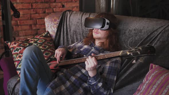 Young Redhead Man Musician Playing Bass Guitar in VR Glasses at Home Sitting on Sofa Slow Motion
