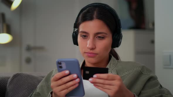 A Young Girl is Sitting on the Couch Wearing Headphones with a Mobile Phone in Her Hands and a Bank