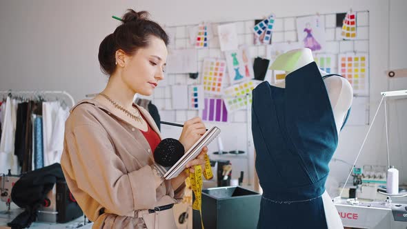 Young Fashion Designer Standing at New Dress Sample on Mannequin and Writing Notes Zoom in Tracking