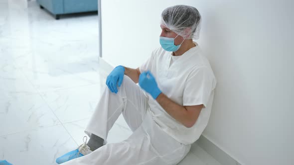 Side View Depressed Man in Medical Uniform and Face Mask Sitting in Corridor Leaning on Wall