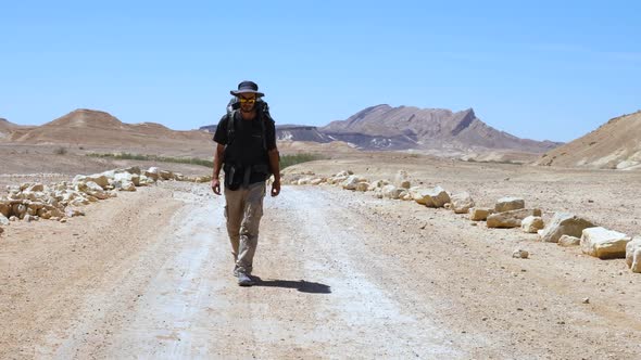 In the heat of the day a man walks down a dusty dirt road in a barren  landscape