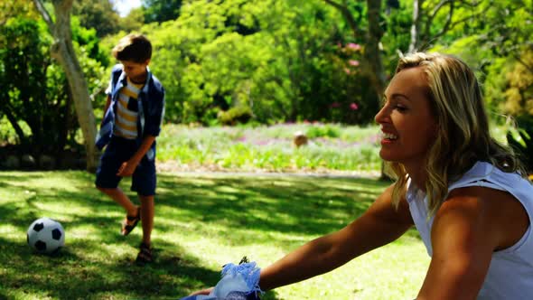 Mother spreading the picnic blanket and son playing football in park