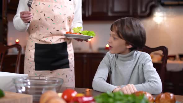 Boy Sitting at Table in Kitchen Waiting for Meal As Unrecognizable Mother Bringing Healthful