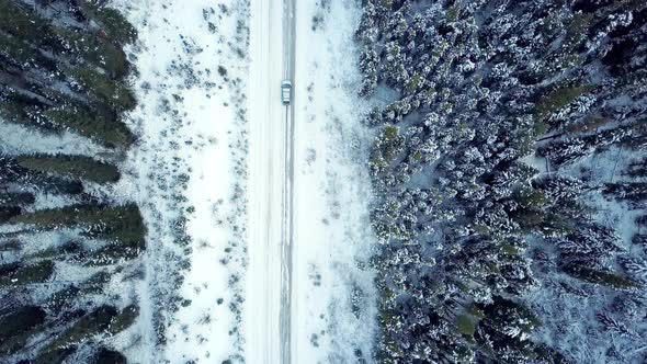 Aerial view looking down as a car drives along a winter road through pine forest