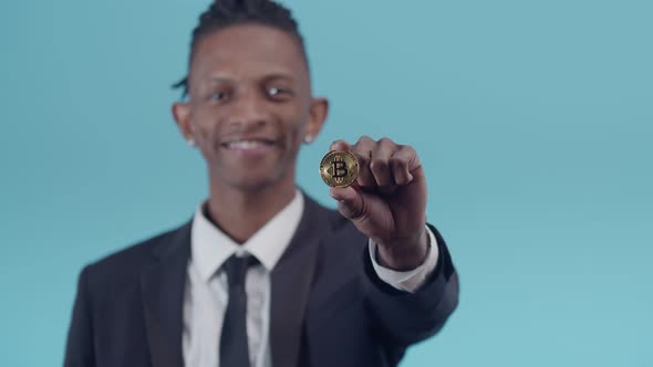 Black Man in an Official Suit Shows a Closeup Bitcoin Coin on the Studio's Blue Background