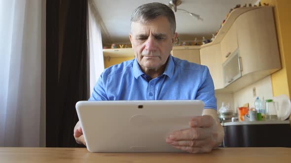Senior Man At The Blue Shirt Sits And Uses A White Tablet Pc At Home