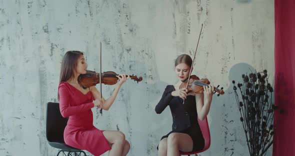 Duet of Female Musicians Sitting on Chairs and Playing the Violins Indoor