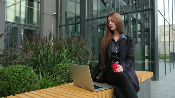 Young Cute Woman with Laptop and Coffee Outdoor on Wooden Urban Bench