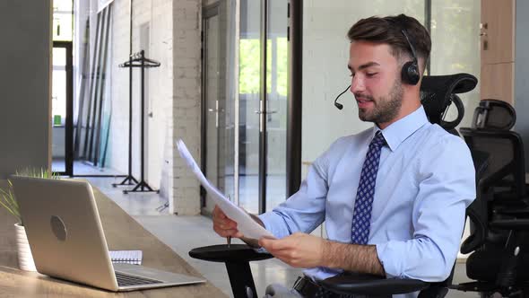 Cheerful young support phone male operator in headset, at workplace while using laptop