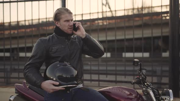 A Young Man Wearing a Leather Jacket Sits on His Motorcycle at the Sunset and Speaks on the Phone