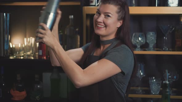 smiling young female bartender shaking cocktail in shaker, medium shot