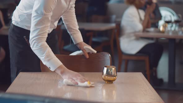 Waiter in Safety Mask Cleaning Table with Disinfectant Spray and Microfiber Cloth in Cafe.