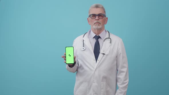 Serious Adult Man Doctor Holds a Phone with an App in the Studio on a Blue Background