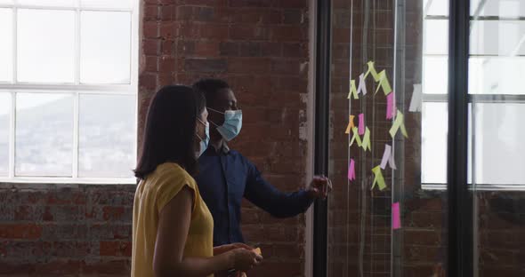 Diverse male and female business colleagues wearing face masks brainstorming pointing to glass wall