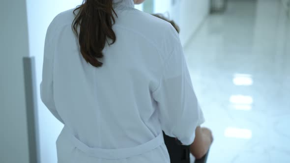 Back View Tracking Shot of Professional Nurse Pushing Wheelchair with Patient Walking on the Left