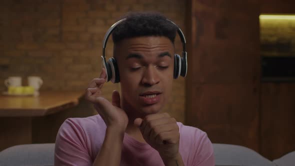 20s African American Man Enjoys Listening the Music in Wireless Headphones Sitting on Sofa at Home