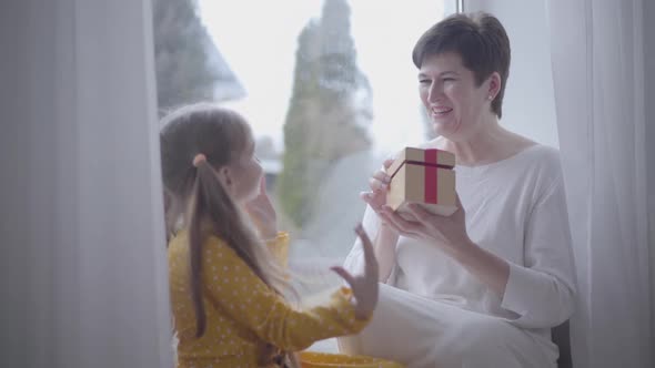 Portrait of Surprised Adult Caucasian Woman Receiving Gift From Cheerful Little Girl. Granddaughter