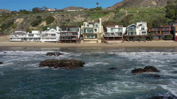 Aerial shot of ocean waves and cliff - waves washing up on a rock in sea and houses in the backgroun