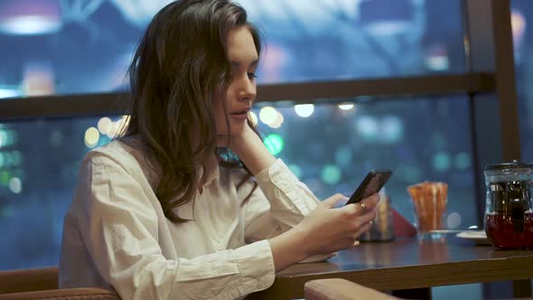 Young Girl Is Texting a Message on the Smartphone While Sitting in a Cafe on the Background of the