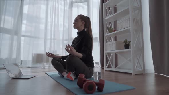 Young Calm Woman Sits in Staff Easy Lotus Pose Near Laptop at Home Interior, Meditating Alone on the