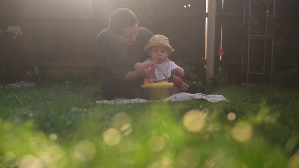 Happy Young Cheerful Mother Holding Baby Eating Fruits On Green Grass