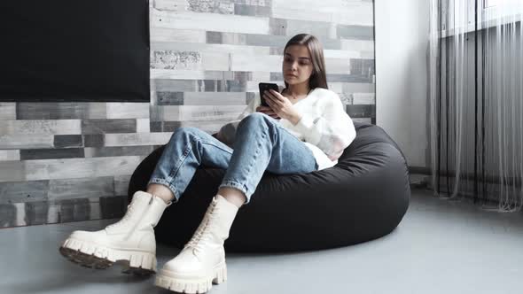 A Girl is on Her Phone Sitting on a Cushioned Chair in a Bright Room