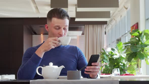 Handsome Happy Man Laughing, Using His Smart Phone at the Cafe During Breakfast