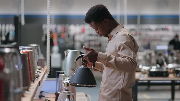 Young Black Man is Comparing Two Electric Kettle in Store Holding Both in Hands and Weighting