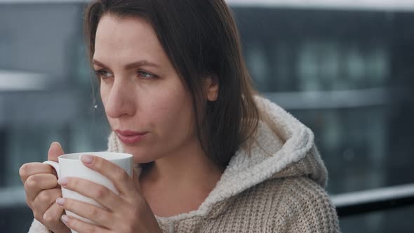 Caucasian Woman Stays on Balcony During Snowfall with Cup of Hot Coffee or Tea