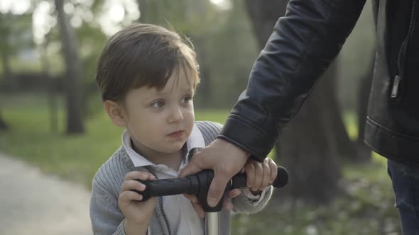 Joyful Caucasian Little Boy Riding Scooter in Sunlight with Young Male Hand Pulling Steering Wheel