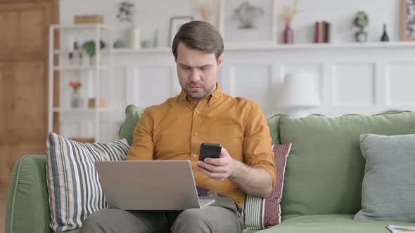 Young Man with Laptop using Smartphone on Sofa